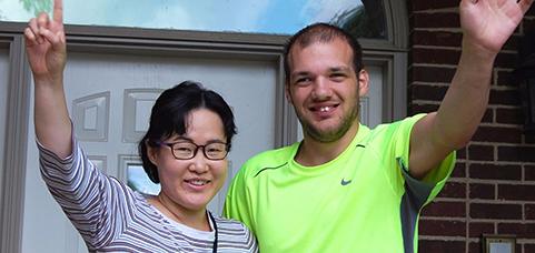 Two smiling people, a woman and a man, raise their arms in celebration in front of a brick house.