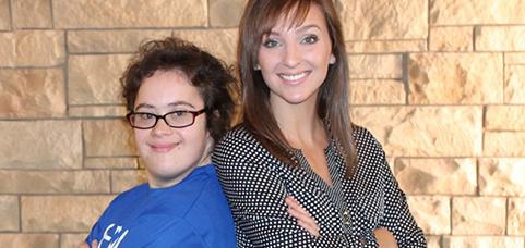 Two people smiling in front of a stone wall, a girl in a blue shirt and a woman with long hair.