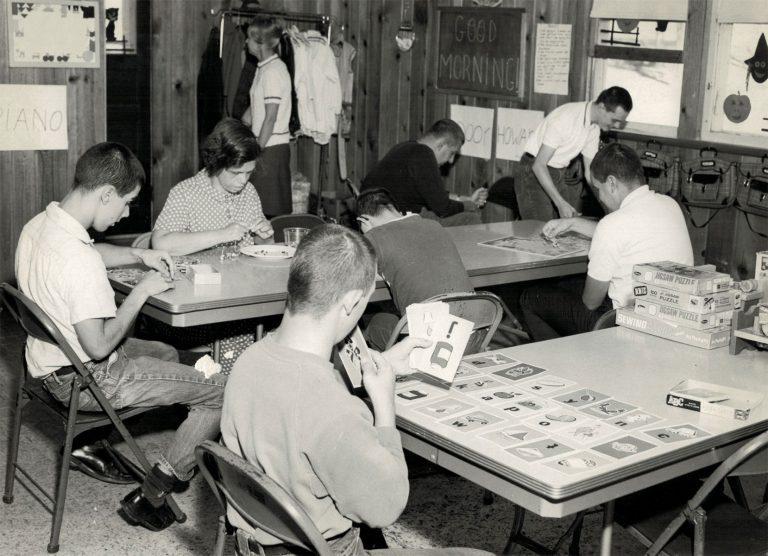 Several men sit around tables in a room, examining papers and playing cards.