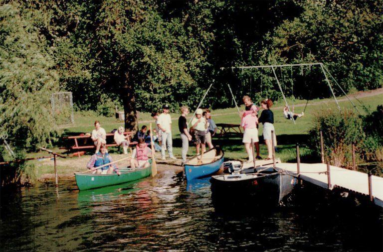 Group of people standing on a wooden dock by a lake with several small boats moored nearby and trees in the background.