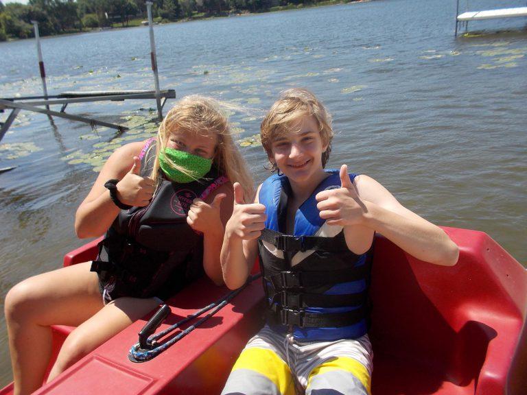 Two children wearing life jackets sit in a red boat on a lake, giving thumbs up.