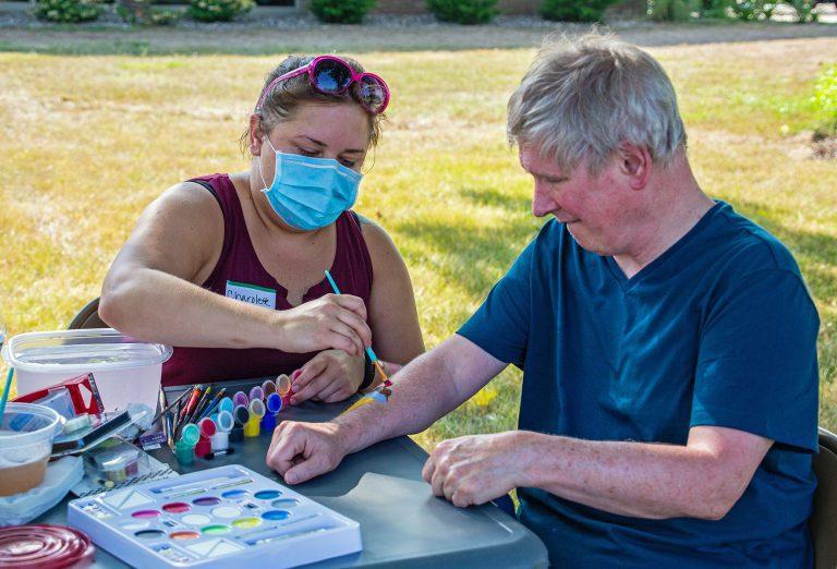 Two people sit at an outdoor table as a woman wearing a blue mask paints a man's nails with nail polish bottles and tools on the table.