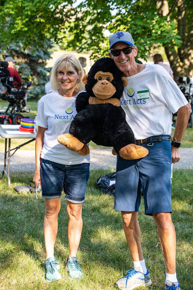 A smiling couple stands on a sunny park lawn, holding a large stuffed teddy bear between them.