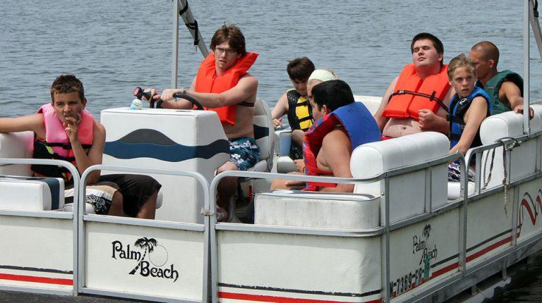 A group of people wearing life jackets sits on a white pontoon boat on calm water.