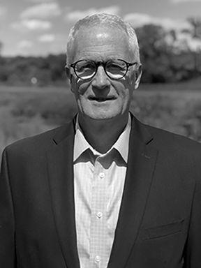 Black-and-white portrait of an older man with gray hair and glasses, wearing a suit jacket and collared shirt outdoors.