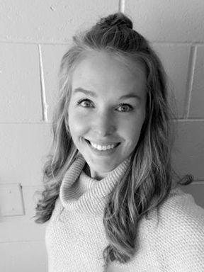Smiling young woman with long wavy hair wearing a light knit sweater, standing against a light brick wall.