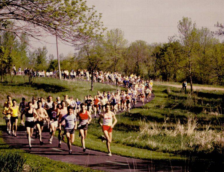 A large group of runners competes in a park road race along a tree-lined path.