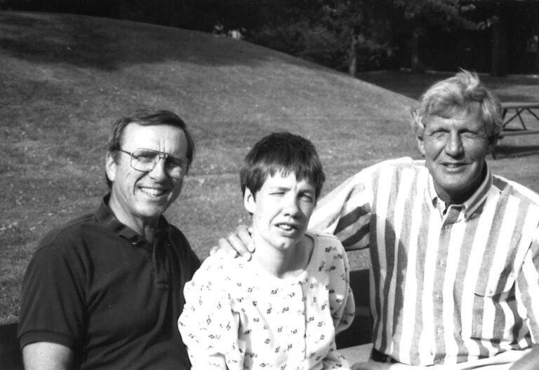 Three people pose outdoors in a park: a man with glasses, a smiling girl, and a man in a striped shirt.
