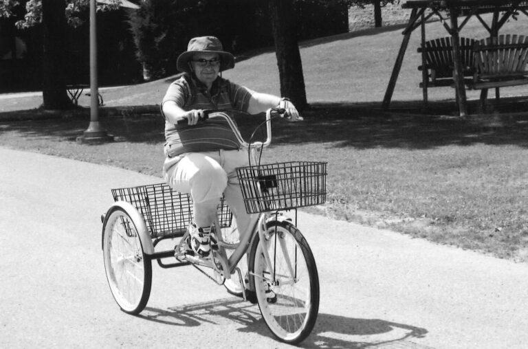 Person wearing a hat rides a bicycle with a front basket along a park path.