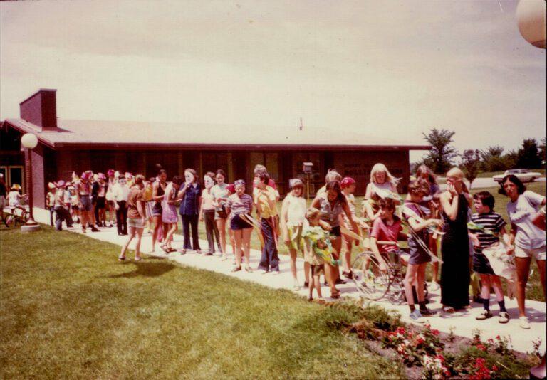 A long line of people, including children, stand along a sidewalk outside a red building with a grassy lawn and flower bed in front.