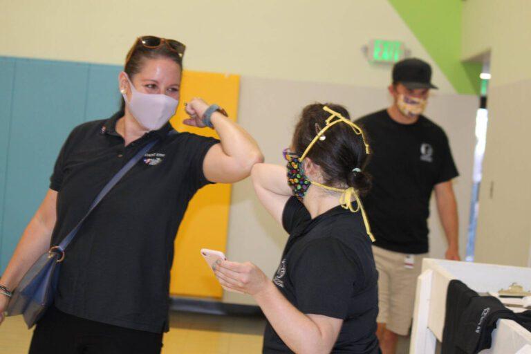 Two women in black shirts pose for a photo in a bright gym, one wearing a face mask, while a man in a cap stands in the background.