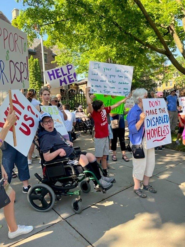 A group of protesters outdoors, including a person in a wheelchair, holding disability-rights signs.