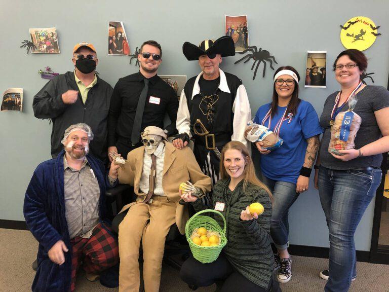 A group of coworkers in Halloween costumes pose together in an office hallway decorated with spider decorations.