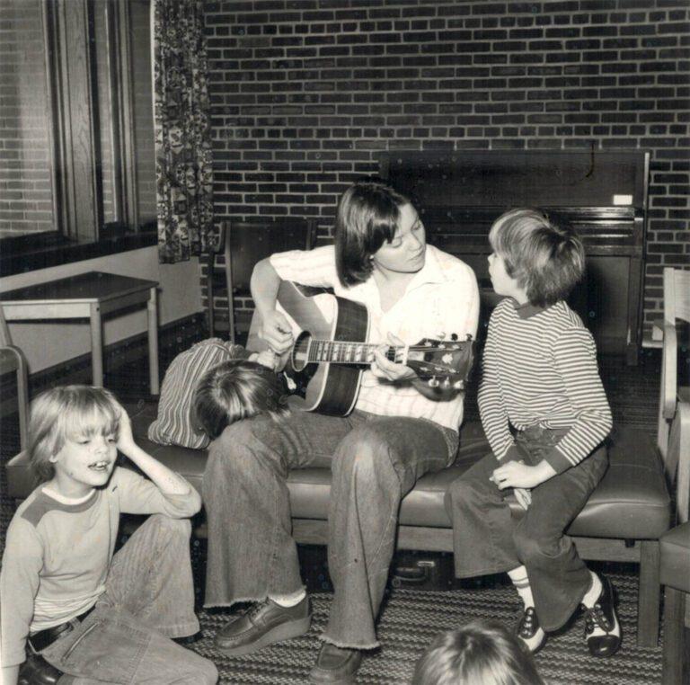 Man sitting on a couch playing an acoustic guitar while several children sit and listen in a living room.