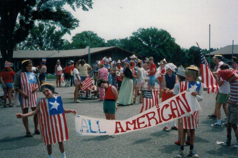 People in a street parade hold a banner reading 'All American' while waving American flags.