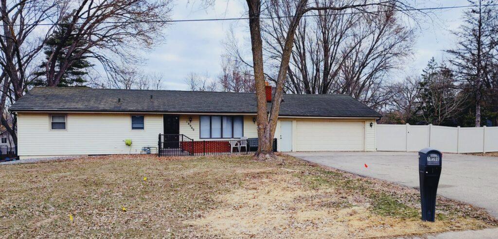 A single-story white suburban house sits behind a leafless tree in a brown front yard, with a driveway on the right and a black mailbox near the street.