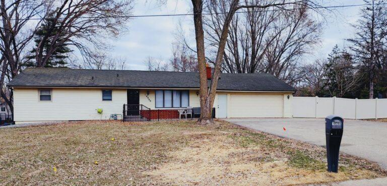 A single-story white suburban house sits behind a leafless tree in a brown front yard, with a driveway on the right and a black mailbox near the street.