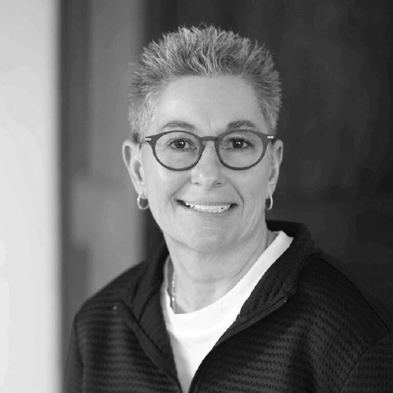 Smiling woman with short gray hair and round glasses, wearing a white shirt and a dark zip-up sweater.