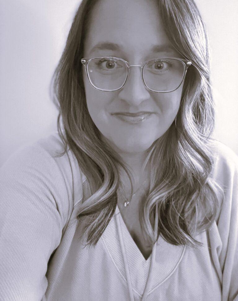 Black-and-white portrait of a woman with glasses and wavy hair, wearing a light top and a necklace, smiling softly at the camera.