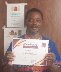 Smiling man in a blue patterned shirt holds a certificate of scholarship in front of a wooden panel background.