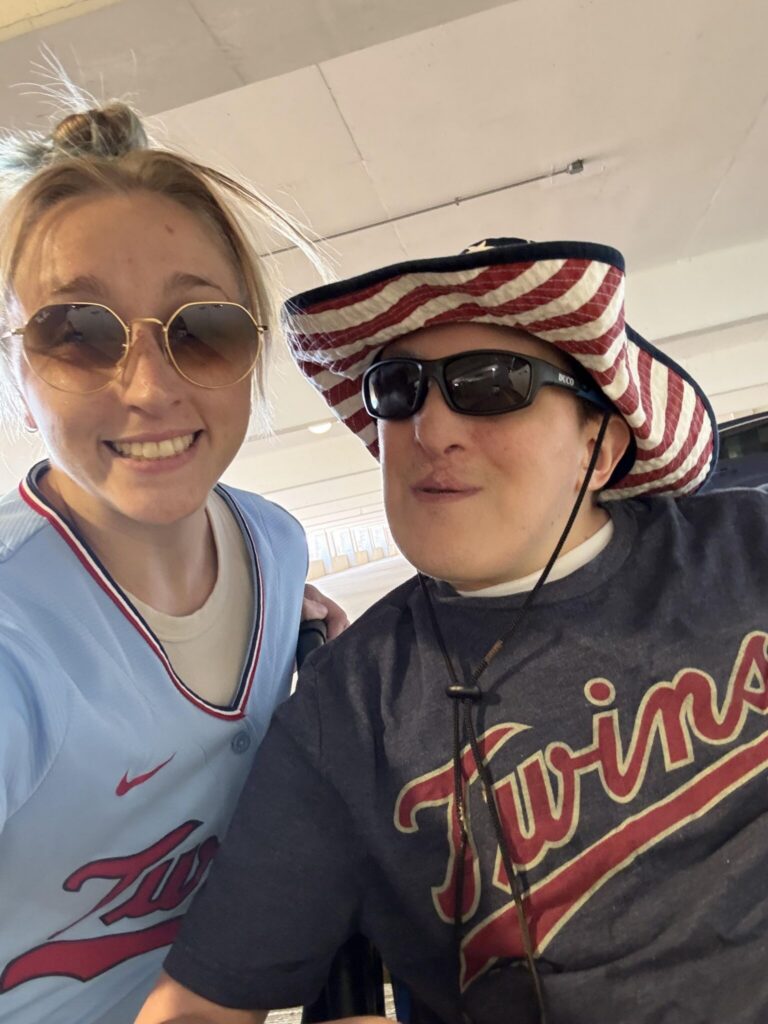 Two people wearing Minnesota Twins apparel pose for a selfie indoors, one in a light blue jersey and the other in a striped red-and-white hat with sunglasses.