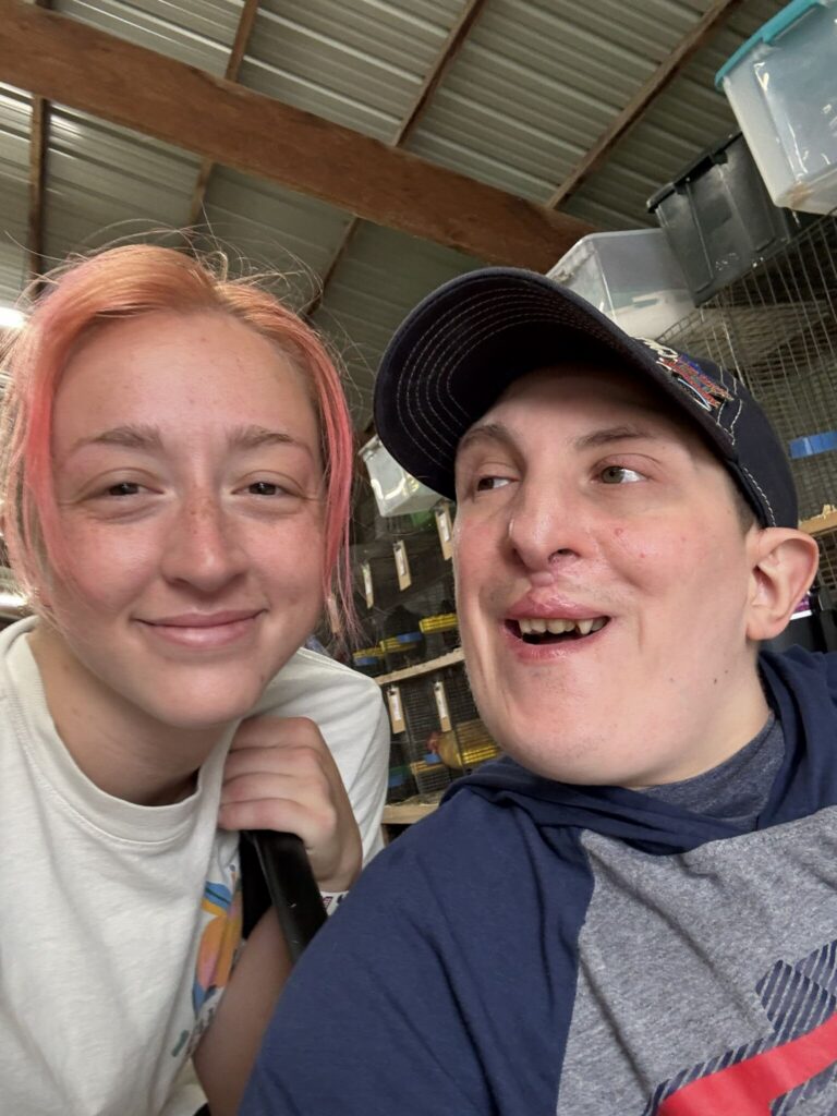 Two people smile at the camera in an indoor, industrial setting—a woman with pink hair and a man wearing a cap.