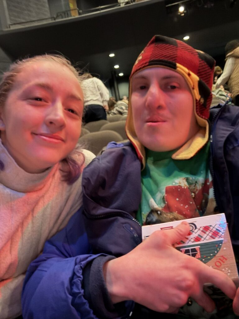 Two people sit in a theater seat smiling at the camera, one wearing a colorful knit hat and the other with light hair, with the person on the right holding a program.