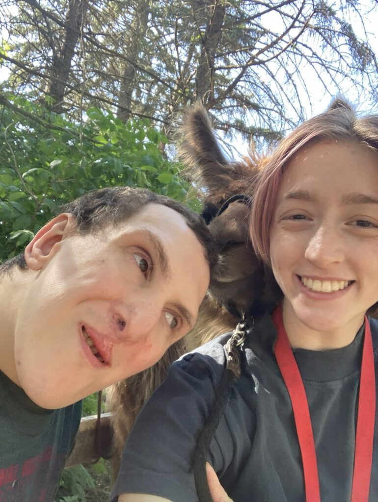 Two people smiling for a selfie with a donkey between them outdoors.