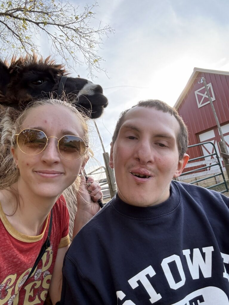 Two friends take a selfie with a dark llama behind them near a red barn on a sunny day.