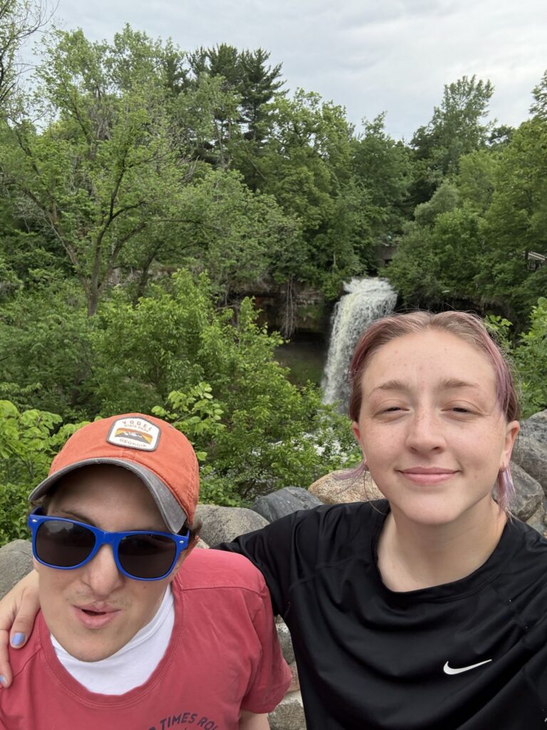 Two friends pose for a selfie on a rocky overlook with trees and a small waterfall in the background.