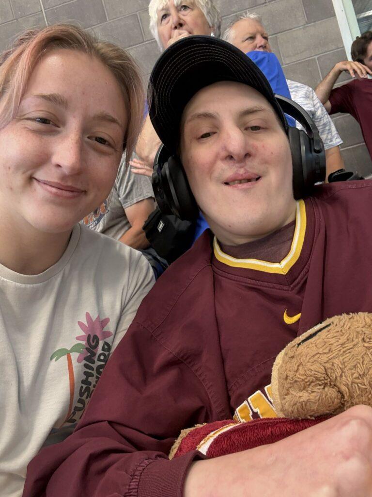 Two smiling people pose for a selfie in stadium seating, with the person on the left in a white shirt and the person on the right in a maroon jersey wearing a helmet and sitting in a wheelchair, holding a teddy bear.