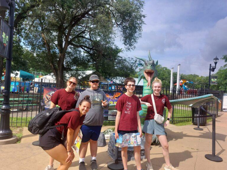 Five friends posing for a photo at a sunny amusement park.