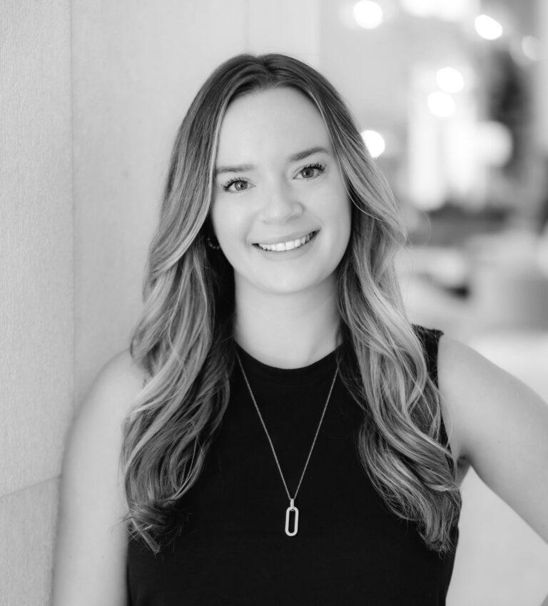 Smiling woman with long wavy hair wearing a sleeveless black top and a pendant necklace, leaning against a wall.