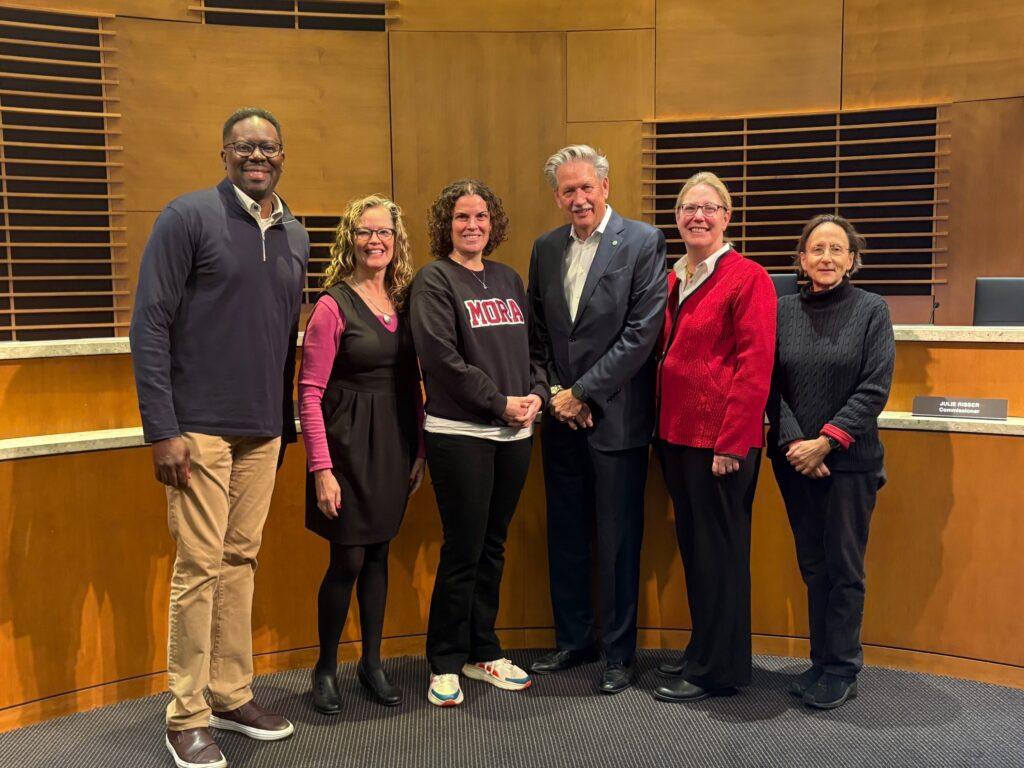 Six adults stand together in a wood-paneled meeting room, posing for a group photo.