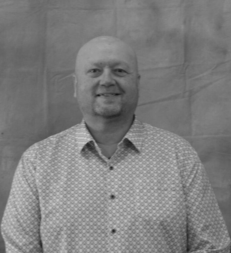 Smiling bald man wearing a patterned button-down shirt stands in front of a plain gray backdrop.