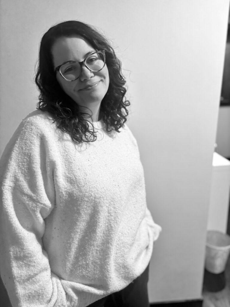 Black-and-white photo of a woman with curly hair and glasses wearing a light sweater, standing indoors and smiling softly.