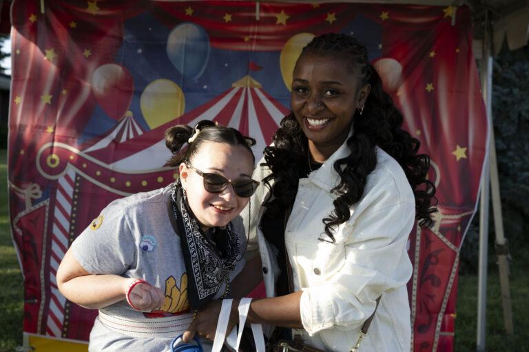 Two people posing and smiling in front of a colorful carnival backdrop with balloons and a striped tent.
