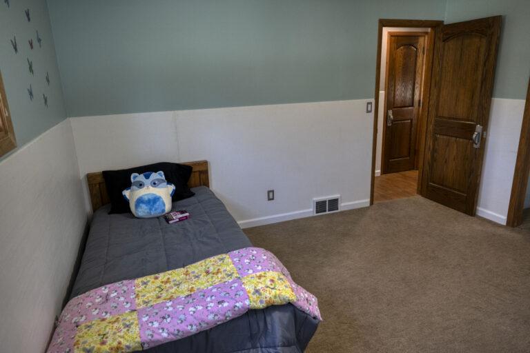 A small bedroom with a single bed along the left wall, a colorful patchwork quilt and a blue stuffed toy on the pillow, beige carpet, two-tone teal and white walls, and an open wooden door on the right.