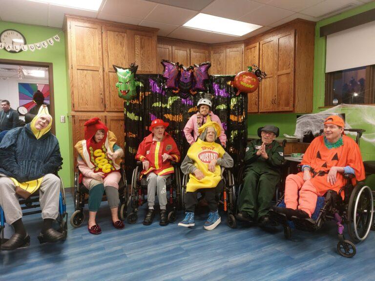 A group of people in colorful costumes, some in wheelchairs, posing together in a room with a decorated backdrop and balloons.
