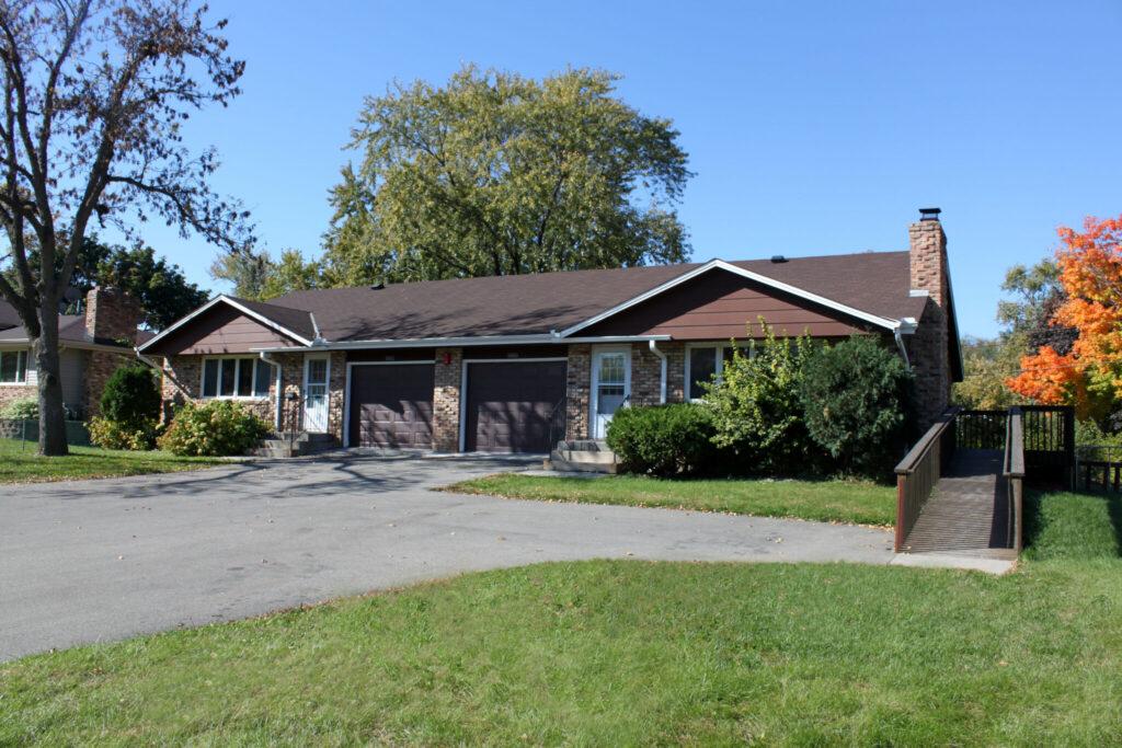 A single-story brick ranch-style house with an attached garage, a chimney, and a well-kept front lawn with trees.