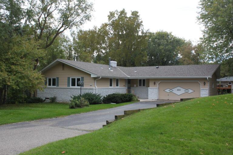 A single-story suburban house with a two-car garage, beige siding, a gray roof, a paved driveway, and a well-kept green lawn with trees in the background.