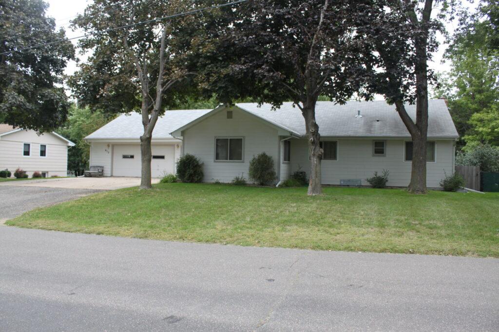 Front view of a single-story suburban house with a two-car garage, a green lawn, and several trees in the front yard.