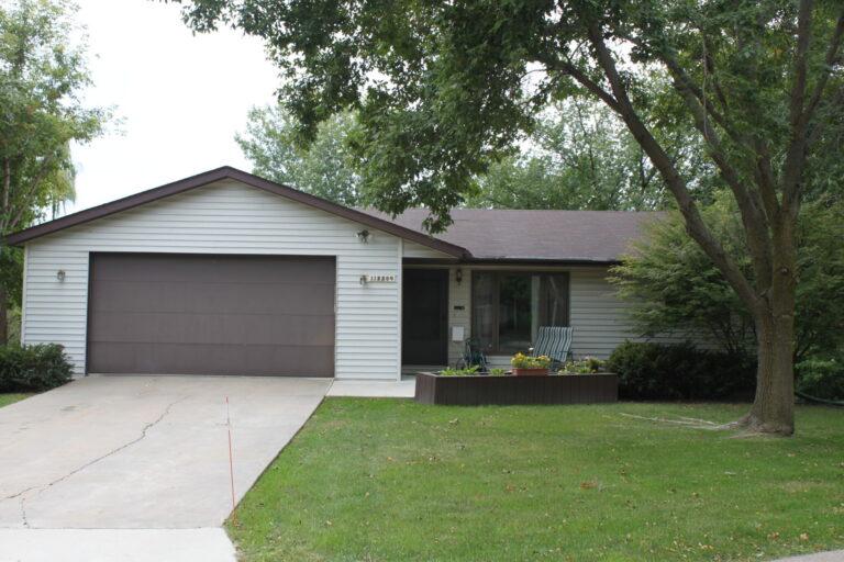 A single-story suburban house with a two-car garage, white siding, a front porch, and a green lawn with a tree on the right.
