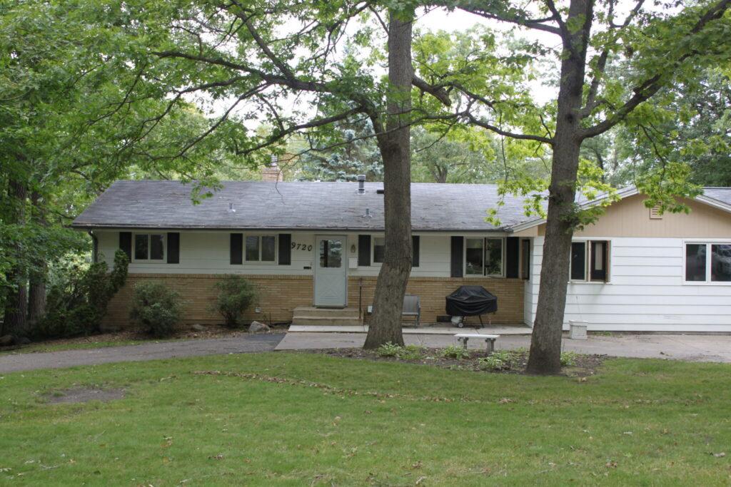 A single-story house with light siding and dark shutters sits behind two large trees, featuring a small front porch and a driveway on the right with a car parked.