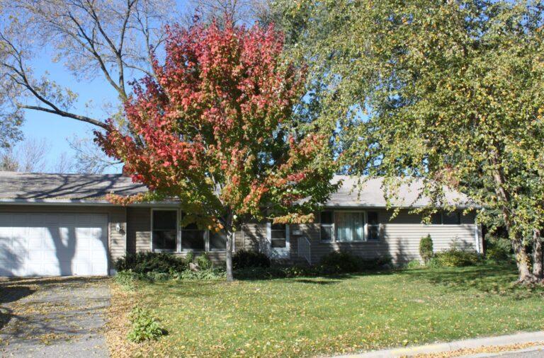 A single-story suburban house with a garage and driveway, in a front yard with a red-leaved tree and other green trees.