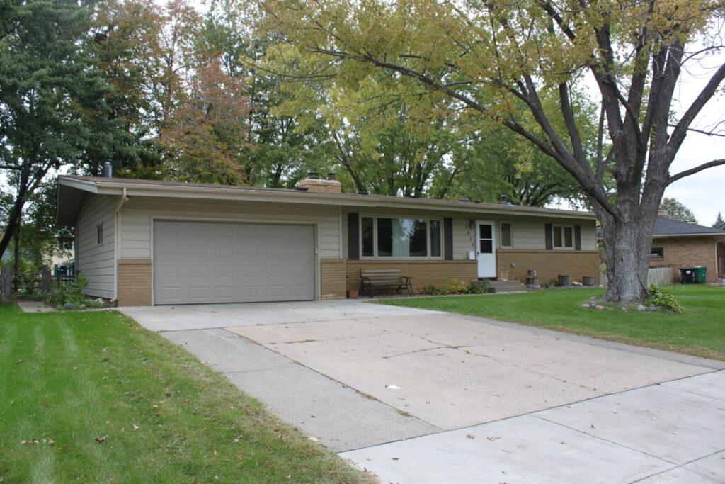 A single-story suburban house with an attached two-car garage, a long concrete driveway, and a green front yard with trees.