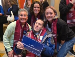 Five friends smile and pose indoors around a man in a wheelchair at the center, who holds a blue certificate.