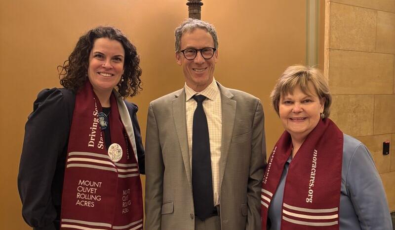 Three adults posing indoors, two women wearing maroon scarves and a man in a gray suit.