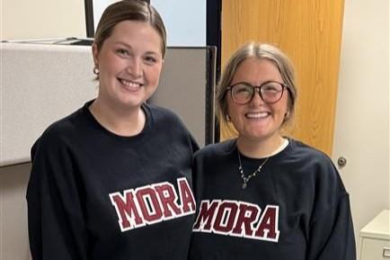 Two smiling women standing indoors, wearing matching black sweatshirts with MORA printed in red on the front.