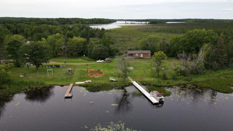 Aerial view of a lakefront property with a wooden dock, a small house, and surrounding trees.